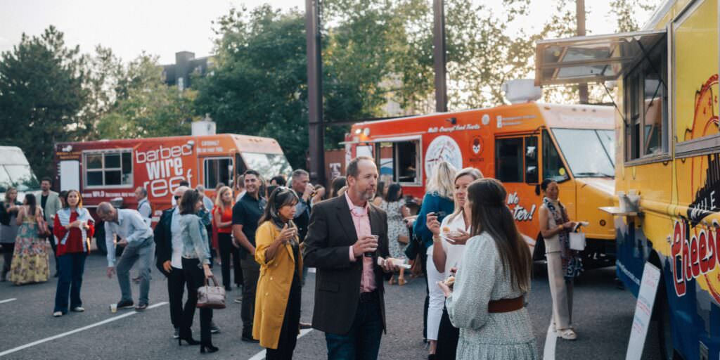 Guests enjoying food truck dining outside during Musana’s Taste to Transform Denver fundraiser, with colorful trucks and lively conversation.